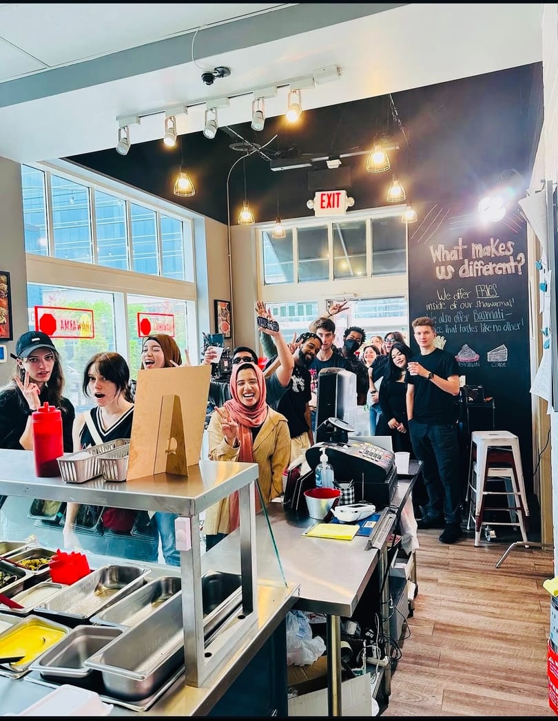 Diverse group of young people smiling and posing behind a restaurant counter with food prep stations.