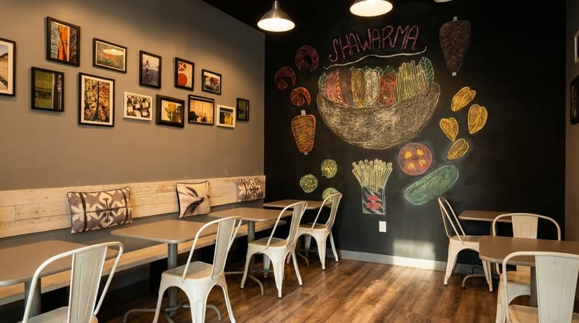 Restaurant interior with a chalkboard mural of shawarma ingredients and framed pictures on the opposite wall.