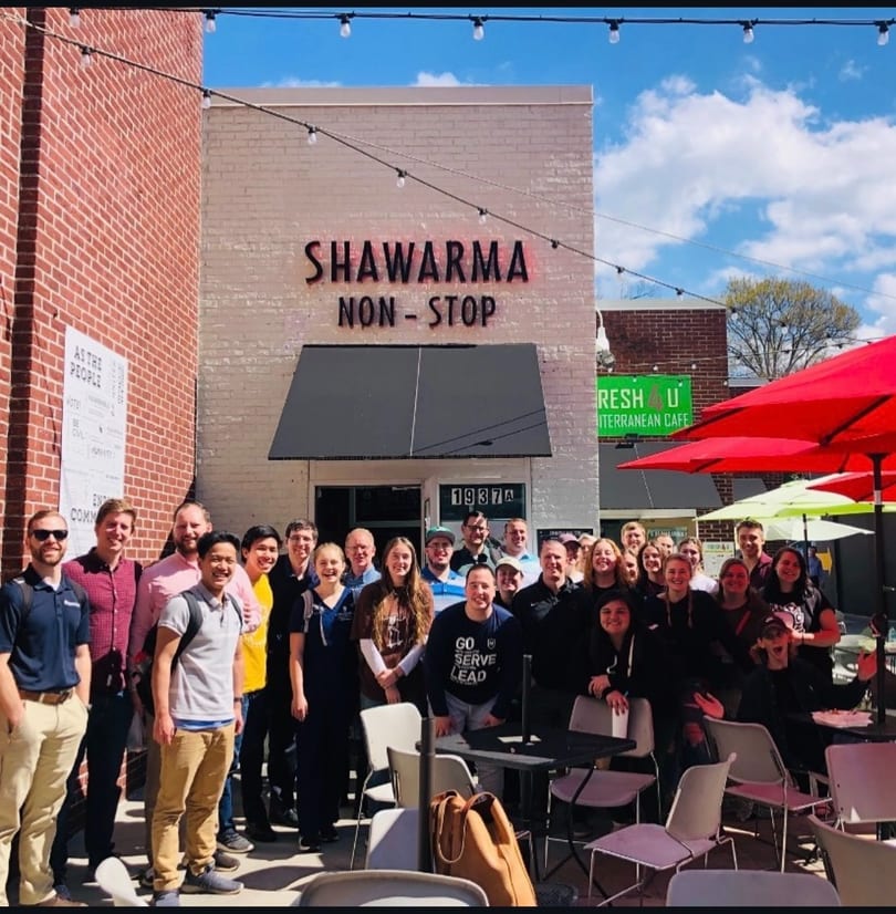 A large group of smiling people gathered outside Shawarma Non-Stop restaurant on a sunny day.