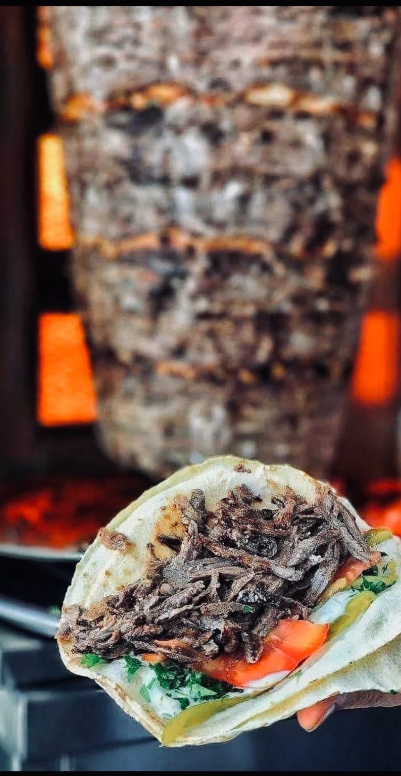 A hand holds a shawarma wrap with meat, lettuce, tomato, and sauce, in front of a meat spit.